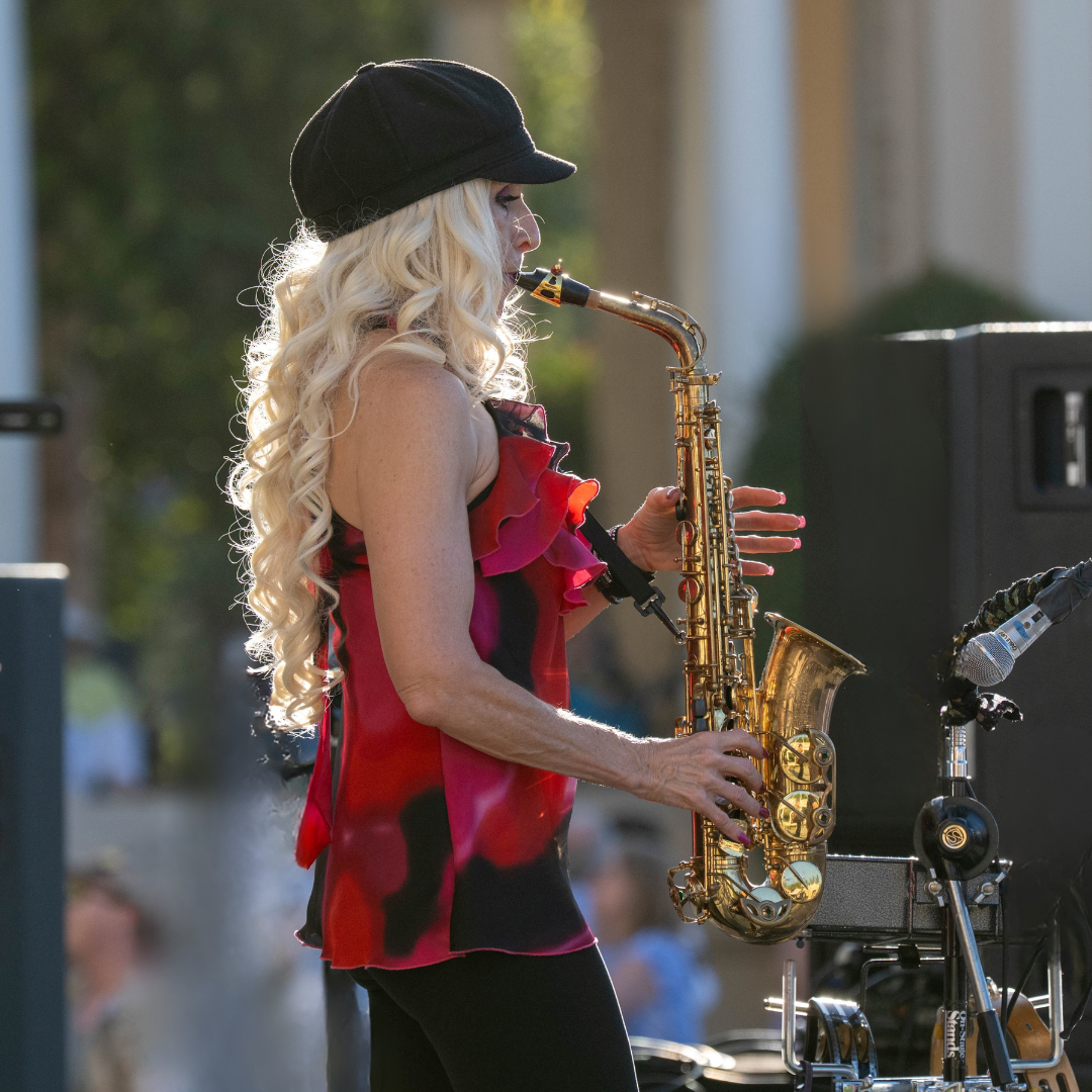 Musician playing saxophone outdoors, wearing a black hat and red top, in side profile.