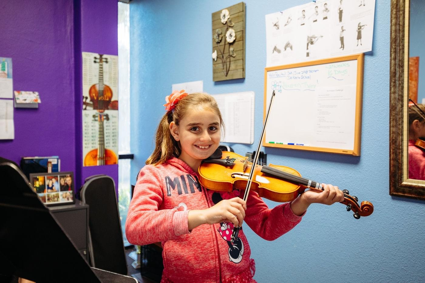 Young girl playing a violin in a music classroom, smiling and wearing a pink sweater.